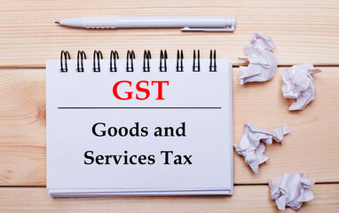 On a wooden background, a white notebook with the inscription GST Goods and Services Tax, a white pen and crumpled white pieces of paper
