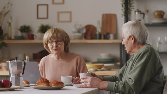 Elderly wife and husband sitting together at kitchen table, having tea, reading news online on laptop and discussing them