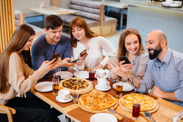 A group of young cheerful friends is sitting in a cafe talking and taking selfies on the phone. Lunch at the pizzeria.