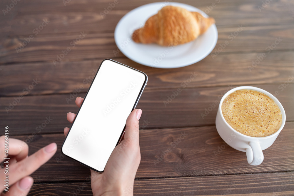 Poster break time. lady holding smartphone with blank screen while drinking coffee and eating croissant at 