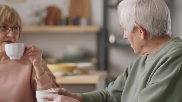 Elderly woman drinking tea, reading news online on laptop screen and chatting with husband in kitchen at home
