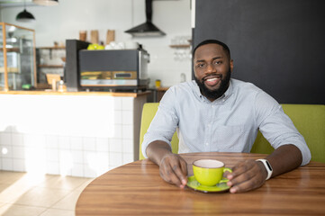Smiling African-American guy is sitting in the cozy cafe and enjoys coffee, a multiracial man rests, has a break, looks at the camera cheerfully