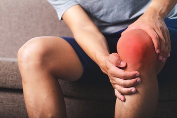 Image of a young man sitting on sofa holding his knees in pain.,Patellar tendon...