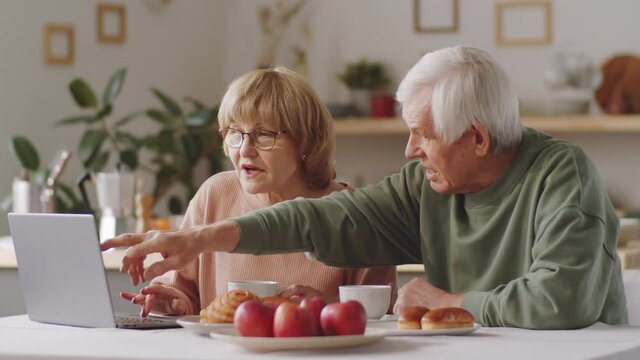 Senior Caucasian couple sitting together at table in kitchen, pointing on laptop screen and discussing online news while having breakfast in morning