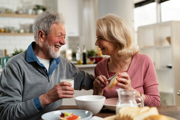 Senior couple eating breakfast in the kitchen. Husband and wife talking and laughing while eating a sandwich...