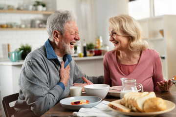 Senior couple eating breakfast in the kitchen. Husband and wife talking and laughing while eating a sandwich...