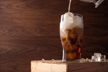 Milk is poured into a glass filled to the top with ice and coffee. A glass of iced coffee sits on a crate against a wooden background