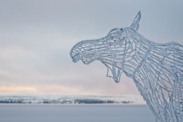 A moose head made of metal bars against the backdrop of the frozen and snow-covered Storsjön Lake in Östersund - 429241303