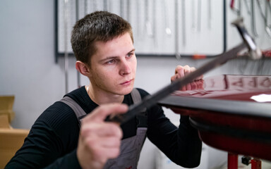 Processing of no paint using dent restoring technology by young technic looking narrowly on leveling surface while he punches it with rubber hammer.