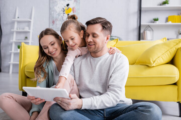 Kid hugging father with digital tablet near smiling mother in living room.