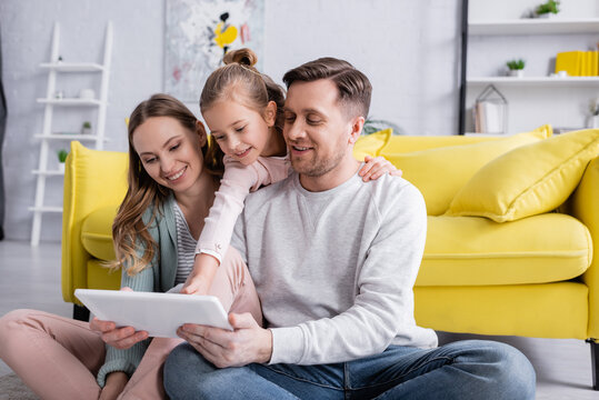 Kid hugging father with digital tablet near smiling mother in living room.
