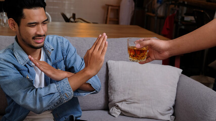 Asian man in blue jean jacket refuses say no and avoid to drink a alcohol whiskey by stopping hand sign male alcoholism concept treatment of alcohol addiction quit booze - obrazy, fototapety, plakaty