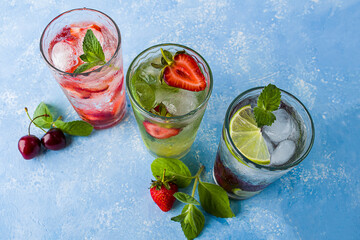 Three glass of refreshing cool detox drink with strawberry, lime, cherry and mint on blue background. Various summer lemonades or ice tea. Mojito cocktails with ice cubes. Healthy eating.