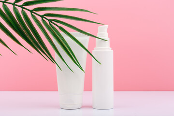 Two white unbranded cream tubes against on white table pink background with palm leaf. Concept of skin care and beauty treatment for smooth, young looking skin