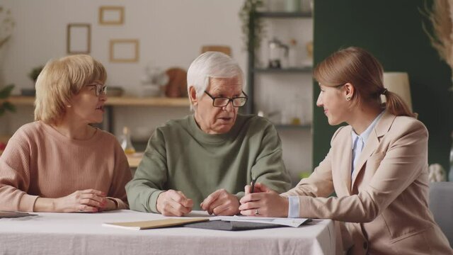 Elderly couple and female financial advisor having discussion, then looking together at camera and smiling while sitting at table at home
