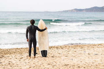 Back view of surfer man wearing diving suit leaning on the surfboard and standing in front of the sea looking the waves