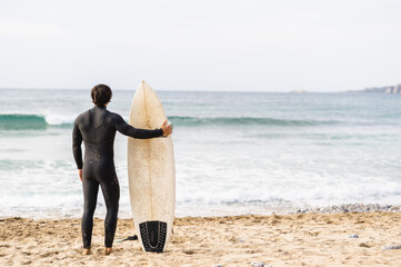 Back view of surfer man wearing diving suit leaning on the surfboard and standing in front of the sea looking the waves