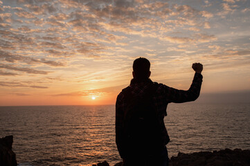 Fototapeta premium Back view silhouette of unrecognizable man standing against the sea looking sunset raising arms and showing victory fist.