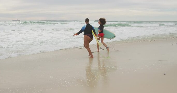 Happy african american female friends running into the sea holding surfboards