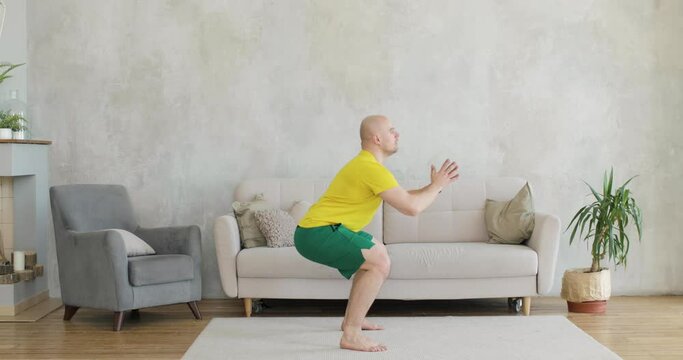 A young man works out doing squats at home. Side view.