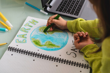 Wide angle view of child girl draws planet earth with wax colors on school notebook for Earth day -...