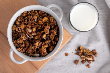 Organic homemade roasted granola cereal with oats, flax, almonds, chocolate in white round bowl with glass of milk on wooden tray or board on linen textile. Top view natural muesli breakfast, flat lay