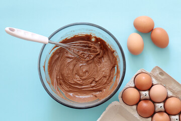 creamy chocolate dough in a bowl with whisk and eggs on blue background