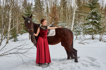 A girl in a vintage red dress stands next to a horse in a winter forest.