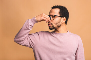 Afro american man isolated against beige background smelling something stinky and disgusting, intolerable smell, holding breath with fingers on nose. Bad smells concept.