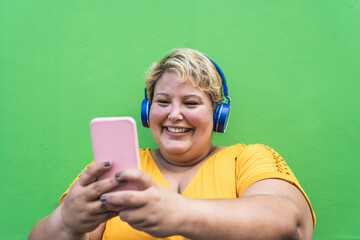 Happy plus size woman taking selfie with mobile smartphone and listening music with wireless headphones - Confident overweight people and technology concept