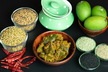 Mango Pickel or Homemade Mango Pickle or aam ka achar or Kairi Loncha in a wodded bowl with Spices and jar and Raw Mango, selective focus isolated on black background