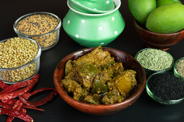 Mango Pickel or Homemade Mango Pickle or aam ka achar or Kairi Loncha in a wodded bowl with Spices, chillies, Raw Mango and jar, selective focus isolated on black background
