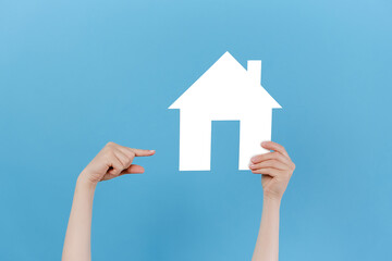 Fototapeta na wymiar Close up of young female hand raising and pointing to small white paper house, posing isolated over blue studio background wall with copy space for advertisement. Property and mortgage concept