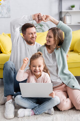 Girl with laptop showing yes gesture near smiling parents on blurred background.