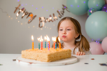 cute little girl blows out candles on a birthday cake at home against a backdrop of balloons....