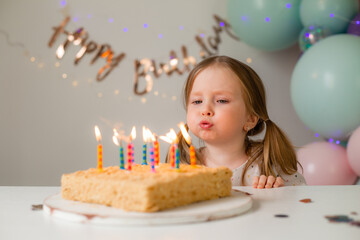 cute little girl blows out candles on a birthday cake at home against a backdrop of balloons....