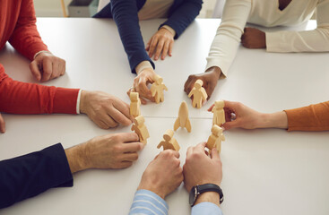 Diverse business team members join wooden human figures around space rocket on table in corporate meeting. Metaphor for teamwork. Symbol of developing new group projects and launching new products - obrazy, fototapety, plakaty