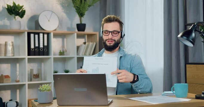 Attractive positive confident skilled young bearded guy in headset discussing report with histogram during video conference with coworkers or clients