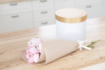 Gold gift box mockup on a wooden table in the kitchen. Glass vase with a bouquet of pink peonies. Scandinavian style interior.