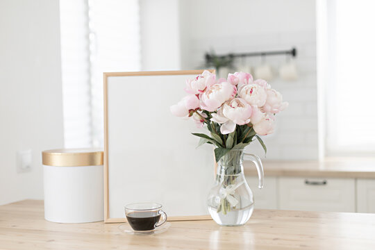 Vertical frame and gift box mockup on a wooden table in the kitchen. Glass vase with a bouquet of pink peonies and a cup of black coffee. Scandinavian style interior.