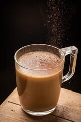 Top view of glass cup of latte coffee, with cocoa falling, on wooden table, black background, in vertical