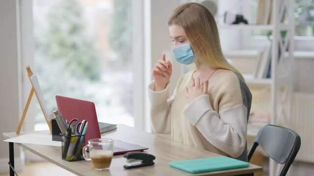 Focused slim young woman in Covid-19 face mask using sign language talking in video chat at home. Portrait of confident Caucasian lady chatting online with deaf person on coronavirus pandemic