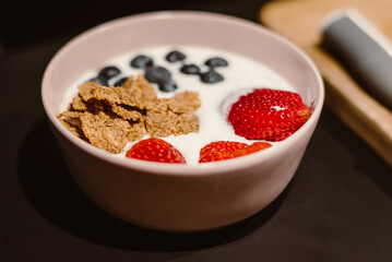 Bowl of oat granola with yogurt, fresh raspberries and blueberries on black wooden board for healthy breakfast.