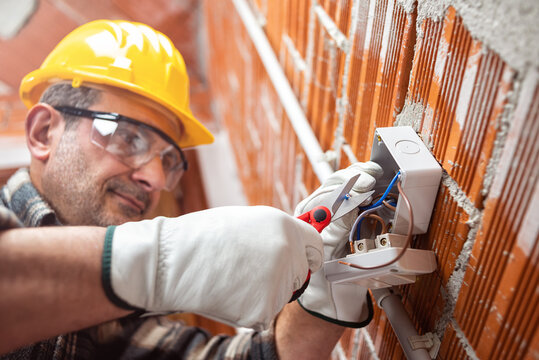 Electrician worker with scissors prepares the electrical cables of an electrical system; wear helmet, gloves and goggles. Workplace safety. 