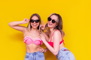 Two young caucasian beautiful happy slim women in swimsuits, denim shorts and sunglasses smile and pose isolated on a bright color yellow background. The girl friends enjoy their summer vacation