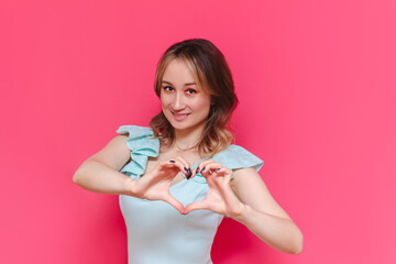A young caucasian pretty lovely smiling brown-haired woman in a light blue dress forming a heart shape with her hands isolated on a bright color pink background. Beautiful cute girl shows her love