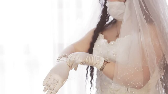 A Jewish bride in a wedding dress and a veil wearing a medical mask puts on elegant white gloves. Medium plan