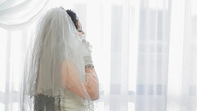 A Jewish bride in a wedding dress and a veil wearing a medical mask in the synagogue hall before the chupah ceremony speaks on a smartphone