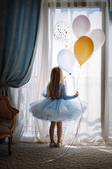 Little Caucasian girl in babydoll dress standing by big french window in bedroom holding baloons in...