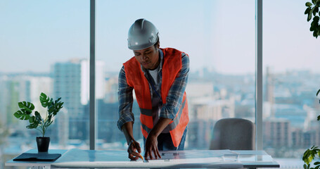 Young african american civil engineer wearing helmet and safety uniform orange vest using mobile phone in office workspace. Architect. Technology. Male portrait.